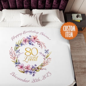 May include: White fleece blanket with a floral wreath design. The wreath has pink and purple flowers and green leaves. In the center of the wreath is the number 80 in gold with the words "Years Loved" below. The text "Happy Birthday Nana! November 20th, 2023" is written in pink below the wreath.