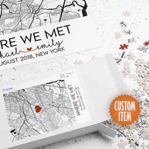 May include: A white jigsaw puzzle box with a black and white map of a city. The box has the text "Where We Met" and "Michael & Emily" with a red heart. The text "03 August 2018, New York" is also on the box. The box is open and the puzzle pieces are scattered around it. The puzzle pieces are white with black outlines. A red heart is on the puzzle piece in the center of the box.