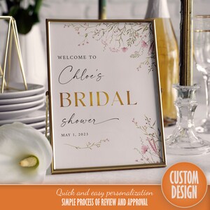 Op de afbeelding: Een gouden lijst met een bloemenontwerp en de tekst "Welkom bij Chloe's Bridal Shower op 1 mei 2023".