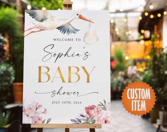 May include: A white sign with a watercolor illustration of a stork carrying a baby bundle. The sign says "Welcome to Sophia's Baby Shower July 30th, 2024".