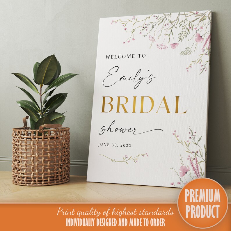 Op de afbeelding: Een wit bruidsdouchebord met een bloemenontwerp en gouden letters. Het bord zegt "Welkom bij Emily's Bridal Shower op 30 juni 2022".