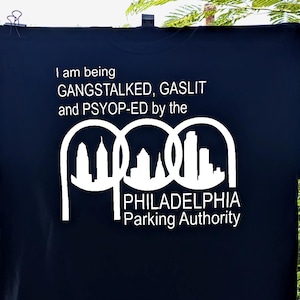 May include: A dark navy blue t-shirt featuring white text that reads: I am being GANGSTALKED, GASLIT and PSYOP-ED by the PHILADELPHIA Parking Authority, accompanied by a white graphic of three interlocking arches containing a city skyline silhouette.