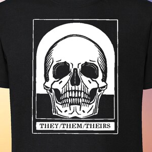 Peut inclure: T-shirt noir avec un motif de crâne blanc à l'intérieur d'un carré blanc. Le texte "THEY/THEM/THEIRS" est situé sous le crâne.
