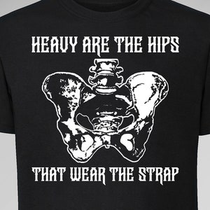 Op de afbeelding: Zwart T-shirt met een witte afbeelding van een bekken en de tekst "HEAVY ARE THE HIPS THAT WEAR THE STRAP." Het shirt heeft ook het Toxic Femme-logo en websiteadres.