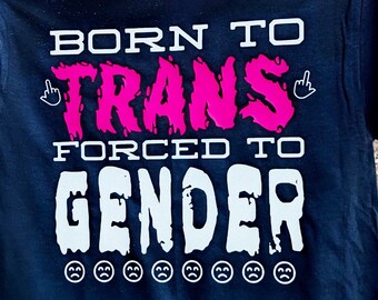 May include: Black t-shirt with the text "Born to Trans Forced to Gender" in pink and white lettering. The text is in a dripping font style. There are eight white sad face emojis below the text.