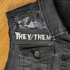 May include: Black denim vest with a patch that says "They/Them" and a safety pin. The vest has a pocket on the front and a button closure.
