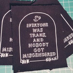 Könnte beinhalten: Schwarzer Stoff-Patch mit weißem Text, der "Jeder war trans, und niemand wurde falsch benannt." lautet. Der Text befindet sich in einem Grabstein-förmigen Umriss mit einem Vogel oben. Der Patch hat einen ausgefransten Rand.