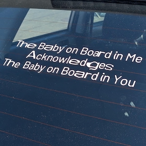May include: White vinyl decal on a car's rear window. The text reads: "The Baby on Board in Me Acknowledges The Baby on Board in You."