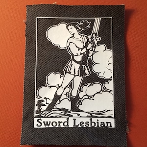 May include: Black and white patch with a woman holding a sword, standing on clouds. The text "Sword Lesbian" is printed below the image.