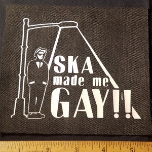 Op de afbeelding: Witte strijk-patch op een zwarte stoffen achtergrond. De patch toont een silhouet van een persoon in pak die onder een lantaarnpaal staat. De tekst luidt "Ska made me gay!"