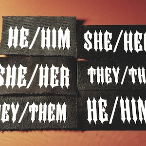 May include: Six black fabric patches with white text. The text on the patches reads "HE/HIM", "SHE/HER", "THEY/THEM".