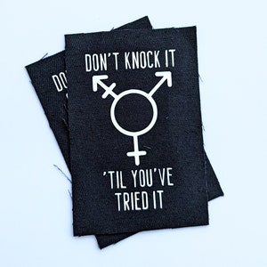 Puede incluir: Parche de tela negro con texto blanco que dice "No lo critiques hasta que lo hayas probado." El parche también presenta un símbolo transgénero blanco.