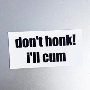 以下が含まれることがあります： 「don't honk! I'll cum」という黒い太字のテキストが印刷された白い長方形のステッカー。ステッカーは明るい灰色の表面に配置されています。