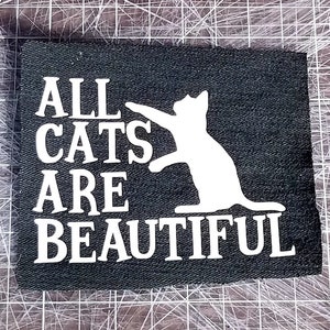 May include: Black fabric patch with white text that reads "All cats are beautiful" and a white silhouette of a cat.