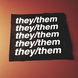 May include: Black fabric patch with the phrase "they/them" repeated five times in white text.