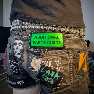 以下が含まれることがあります： パッチとスタッズベルトが付いた濃いグレーのデニムショーツ。パッチには、スカル、スケルトン、「TOXIC LAVA」の文字が含まれています。緑色のパッチには「ADDITIONAL PARTS INSIDE」と書かれています。