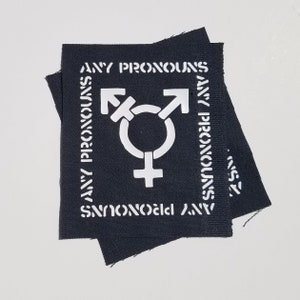 May include: Black fabric patch with white text that reads "ANY PRONOUNS" and a white transgender symbol in the centre.