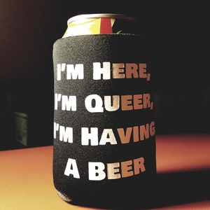 May include: A black can cooler with the text "I'm here, I'm queer, I'm having a beer" printed in white.