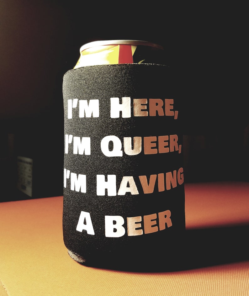 May include: A black can cooler with the text "I'm here, I'm queer, I'm having a beer" printed in white.
