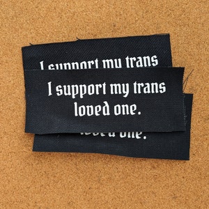 May include: Black fabric patches with white text that reads "I support my trans loved one." The patches are rectangular and stacked on a brown surface. The text is in a gothic font.
