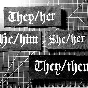 May include: Four black fabric patches with white text in a gothic font. The patches read "They/her", "He/him", "She/her", and "They/them".