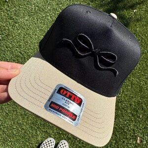 May include: Black and beige baseball cap with a black bow on the front. The cap has a mid profile and is labeled "OTTO" with the text "Classic Fit OSFM" and "Most Popular Fit Stylish Look Premium Fabric".