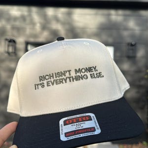 May include: A beige and black baseball cap with the text "RICH ISN'T MONEY. IT'S EVERYTHING ELSE." The cap has a curved brim and a black button on top. The brand label reads "OTTO CLASSIC OSFM MID PROFILE MAXIMUM COMFORT".