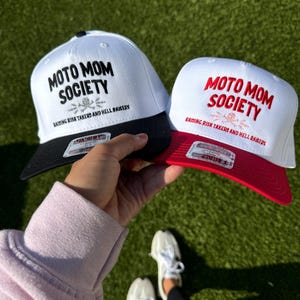 May include: Two white baseball caps with black bills. The caps have black embroidery that reads "Moto Mom Society" and "Raising Risk Takers and Hell Raisers". One cap has a black bill and the other has a red bill.