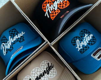May include: Four trucker hats in a cardboard box. The hats are black, blue, beige, and light blue. Each hat has a checkered pattern and a white cursive word on the front. The words are "Los Angeles", "Laster", "Suganski", and "Dishaw".