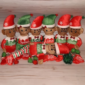 Può includere: Un gruppo di sei bambole di peluche a forma di elfo con la pelle marrone, che indossano abiti rossi e verdi. Ogni elfo ha un nome ricamato sul colletto e l'anno "2020" sulla sciarpa o sulla gamba. Un elfo è sdraiato.