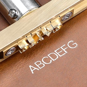 May include: A brass branding iron tool with a long silver-toned metal handle, featuring a horizontal bar that holds interchangeable metal letter stamps. The tool is positioned above a brown leather surface with the letters ABCDEFG embossed.