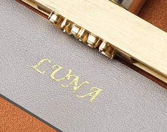 May include: A brass metal branding iron tool with a threaded rod and a silver-toned cylindrical handle, positioned over a rectangular gray leather strip. The tool features a metal stamp that has embossed the word LUNA in gold on the leather surface.