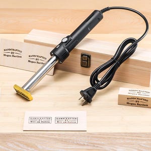 以下が含まれることがあります： 黒いハンドルと黄色の先端が付いた木材焼入れツール。木箱といくつかの木製ブロックの隣にあります。ブロックと箱には「HANDCRAFTED BY Megan Bartlett」と「HANDCRAFTED BY Will'am Duirin」の文字が刻まれています。