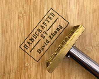 Op de afbeelding: Close-up van een houten oppervlak met de tekst "HANDCRAFTED BY David Zhang" in zwart gestempeld. Een messing brandijzer met een zilveren handvat staat op de voorgrond, klaar om het hout te stempelen.