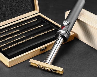 May include: A black electric branding iron with a temperature dial and a metal heating element attached to a brass stamp head. The tool is positioned next to a light wood storage box containing rows of metal character inserts for customization.