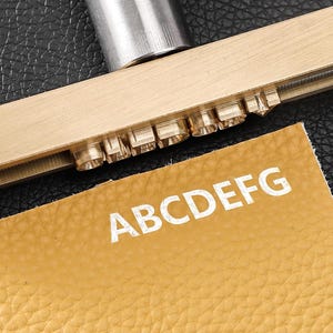 May include: A brass metal stamping tool with a linear arrangement of raised letter characters, positioned above a piece of tan textured leather that displays the embossed white letters ABCDEFG, showing the result of the stamping process.