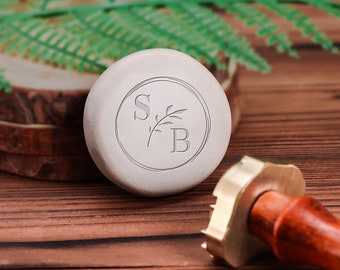 May include: A white round wax seal stamp with the initials "S.B." and a small branch design inside a circle. The stamp is on a wooden surface next to a brass and wooden handle.