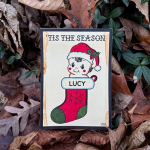 May include: A Christmas-themed illustration featuring a baby wearing a Santa hat and a red stocking with the name "LUCY." The text "'TIS THE SEASON" is at the top. The artwork has a vintage style with a black border and is set against a background of autumn leaves.