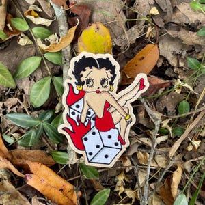 May include: Embroidered patch featuring a cartoon woman in a red dress, perched atop a pair of dice engulfed in flames. The patch has a beige border, black hair, and yellow earrings. The dice are white with black dots.