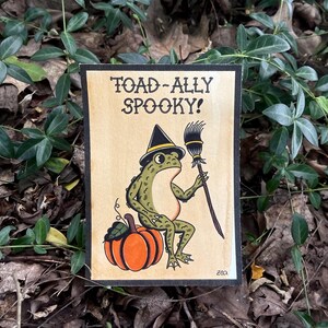 May include: A Halloween-themed illustration featuring a green frog wearing a witch's hat and holding a broom. The frog sits on an orange pumpkin. The text "TOAD-ALLY SPOOKY!" is at the top of the image, with a black border.