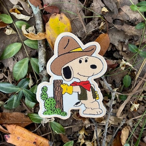 May include: Embroidered patch featuring Snoopy in a cowboy hat, red scarf, and yellow gloves, leaning against a wooden fence with a cactus. The patch has a white border and is set against a backdrop of leaves and foliage.