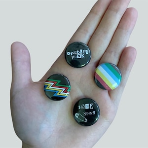 May include: Four round black button pins with different designs. The top pin says "Disabled Punk" in white letters. The bottom pin says "Safe Space" in white letters with a safety pin graphic. The left pin has a colorful zigzag pattern. The right pin has a multi-colored striped design.