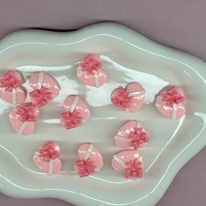 May include: A collection of pink heart-shaped gift boxes with white ribbon and pink bows, arranged on a white, organically shaped ceramic dish. The boxes are glossy and appear to be made of resin or a similar material. The background is a solid, muted purple.