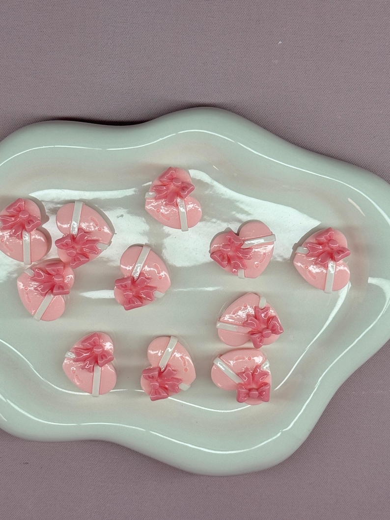 May include: A collection of pink heart-shaped gift boxes with white ribbon and pink bows, arranged on a white, organically shaped ceramic dish. The boxes are glossy and appear to be made of resin or a similar material. The background is a solid, muted purple.