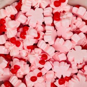 May include: A close-up shot of a pile of pink, flower-shaped candies with red cherry-like accents. The candies are glossy and appear to be made of a hard candy or gummy material. The image is brightly lit, showcasing the details of each piece.
