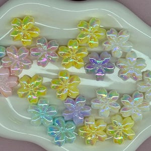 May include: A collection of iridescent flower-shaped embellishments in various pastel colors, including yellow, pink, purple, and blue. The decorative pieces are arranged on a white, scalloped-edge dish, creating a visually appealing display.