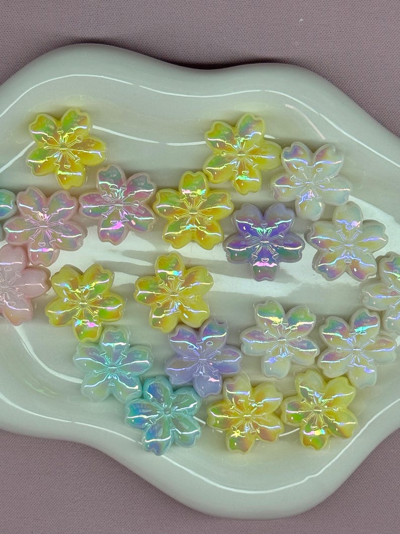 May include: A collection of iridescent flower-shaped embellishments in various pastel colors, including yellow, pink, purple, and blue. The decorative pieces are arranged on a white, scalloped-edge dish, creating a visually appealing display.