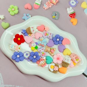 May include: A collection of colorful resin embellishments in various shapes, including flowers, hearts, stars, and cartoon animals. The embellishments are in a white, scalloped dish, with a pink background.