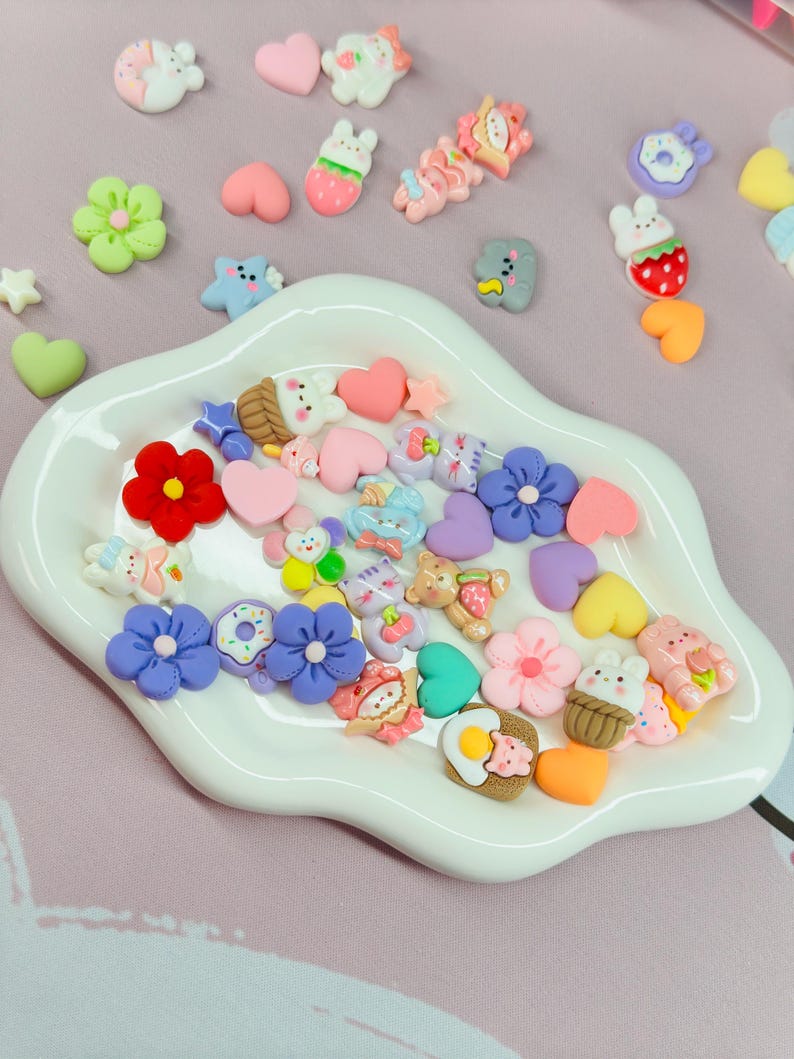 May include: A collection of colorful resin embellishments in various shapes, including flowers, hearts, stars, and cartoon animals. The embellishments are in a white, scalloped dish, with a pink background.
