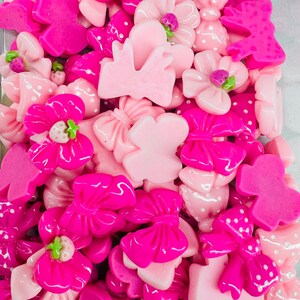 May include: A collection of pink and fuchsia resin embellishments. The assortment includes flowers, bows, and dress shapes. Some flowers have green accents, and some bows have white polka dots. The pieces vary in shade from light pink to bright fuchsia.